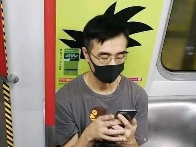 Muž sediaci v metre s maskou na tvári, použil mobil, jeho účes sa zdanlivo spája s obrázkom v pozadí.
