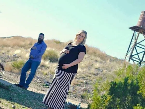 Na piesočnom kopci tehotná žena a stojaci muž pózujú v prírodnej scenérii s vodnou vežou na pozadí.