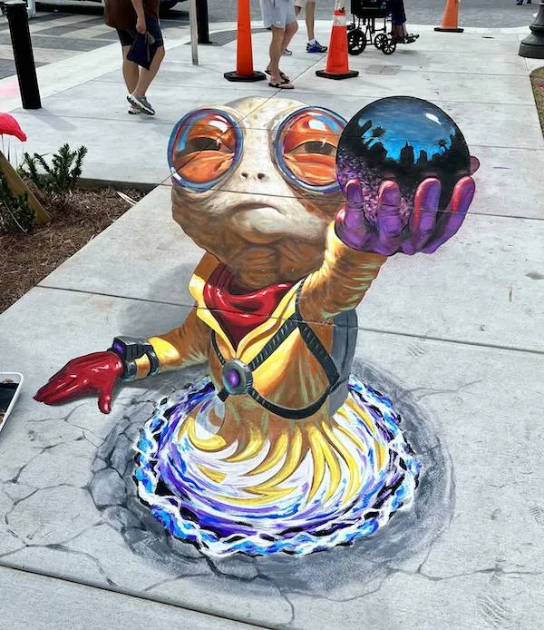 Kreatívna 3D kresba mimozemšťana na chodníku držiaceho kryštálovú guľu s odrazom mesta.