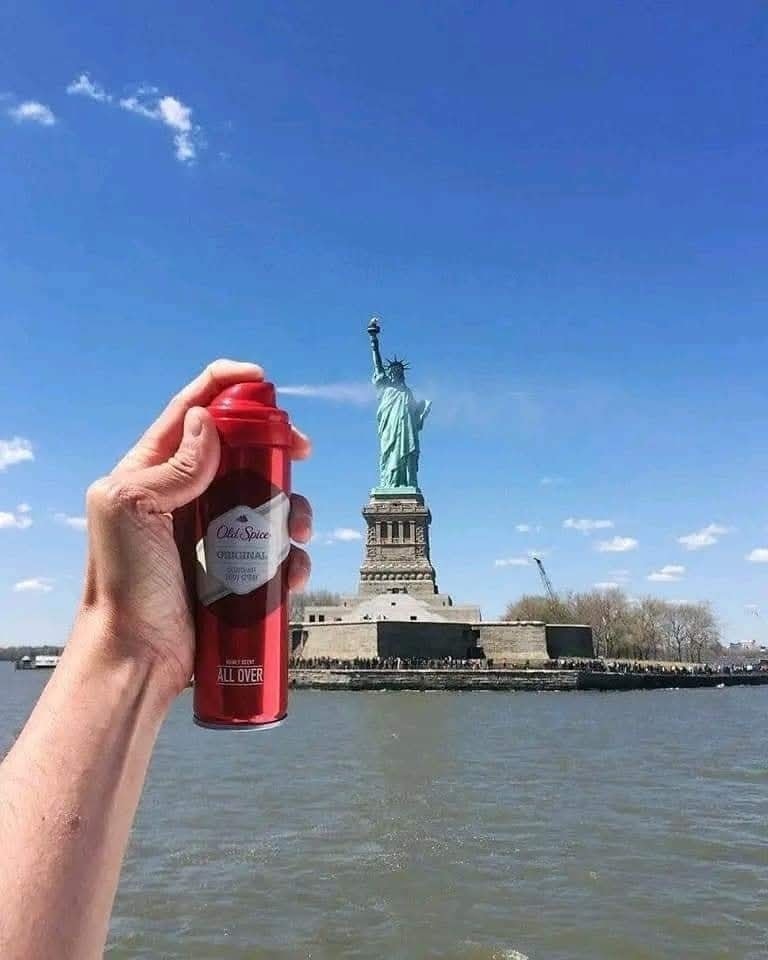 Ruka drží červený sprej pred Sochou slobody, vtipná optická ilúzia na slnečnej oblohe.