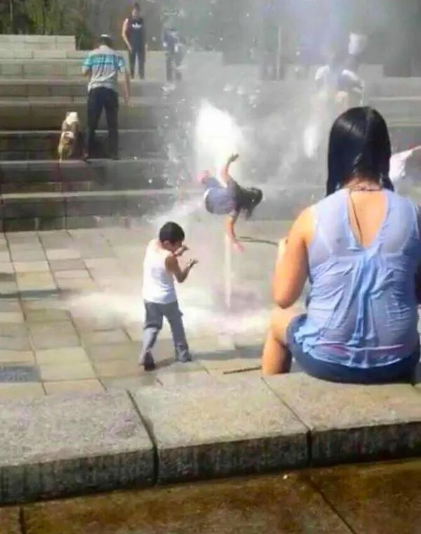 Osoba pri fontáne s vodotryskom skáče do vzduchu, dieťa fotoaparátom zachytáva moment. Letný deň plný zábavy.