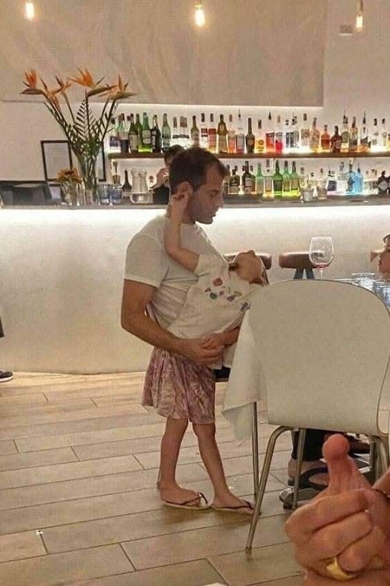 Otec drží spiace dieťa v náručí v reštaurácii pri stole s pohárom vína.