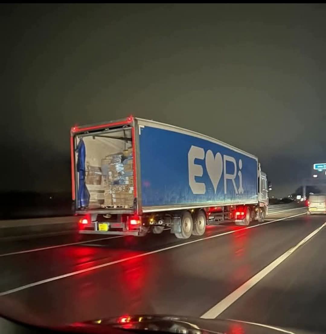 Kamión s otvoreným nákladovým priestorom na nočnej diaľnici, modrá plachta s nápisom a symbolom srdca.