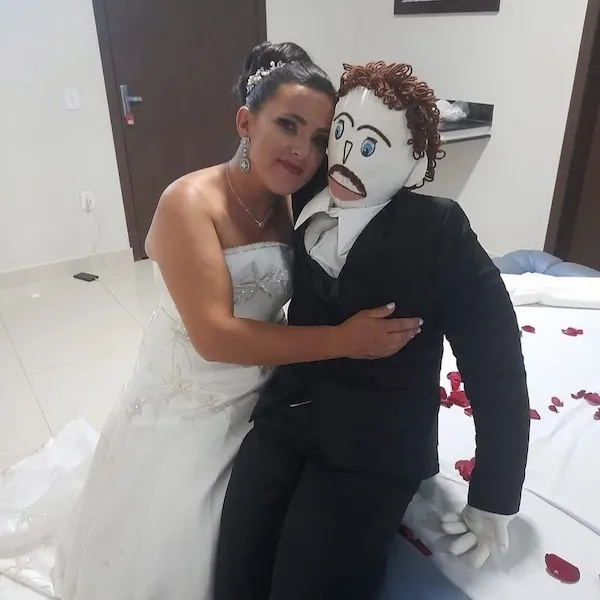 Nevesta objíma figurínu ženicha v izbe so svadobnými dekoráciami a ružovými lupeňmi na posteli.