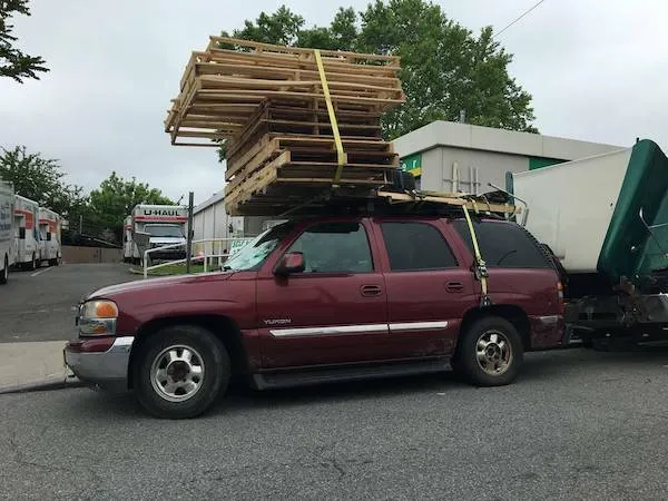 Červené SUV s veľkým nákladom drevených paliet na streche, zaparkované na ulici pri priemyselnej budove.