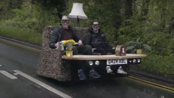 Dvaja ľudia na gaučovom vozidle s lampou a kvetmi na ceste v lese, s úsmevmi a ochrannými okuliarmi.