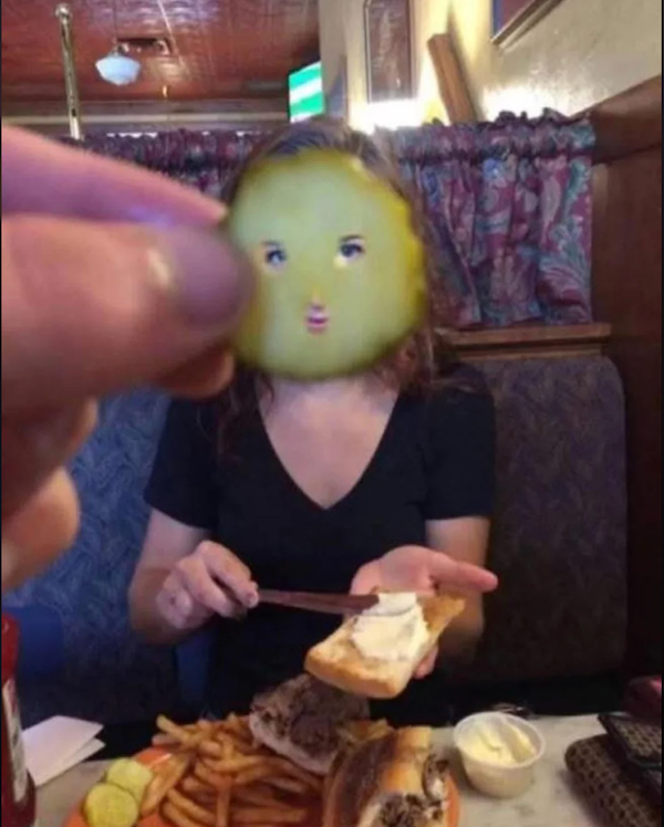 Osoba držiaca kyslú uhorku, ktorá vytvára ilúziu tváre pri príprave sendviča v reštaurácii.