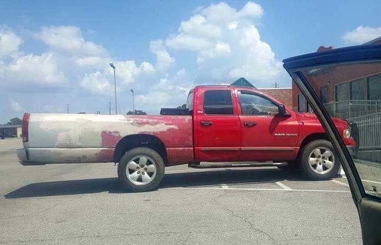 Červený pickup s dlhou karosériou a viditeľnými opravami na parkovisku pod jasnou oblohou.