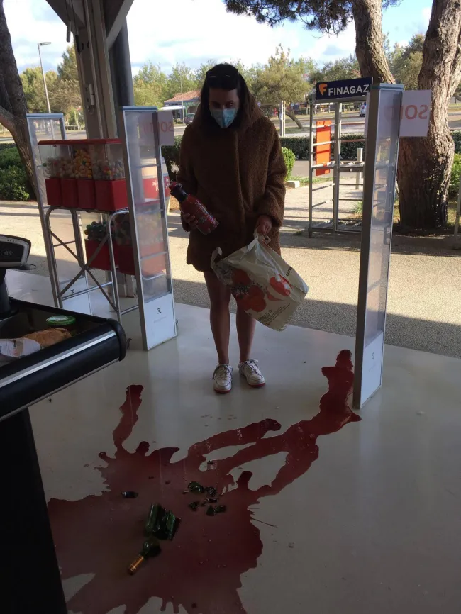 Osoba s rozliatou červenou tekutinou a rozbitou fľašou v obchode, drží tašku a chráni sa rúškom.