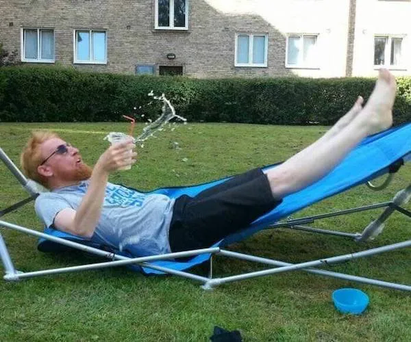 Muž povyšľahol na hojdacej sieti, nápoj sa mu rozlieva, zatiaľ čo relaxuje na trávniku pred budovou.