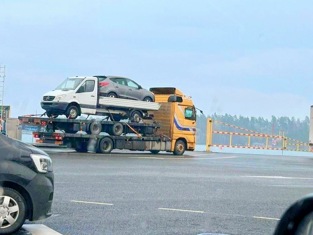 Kamión prepravujúci dva autá na viacúrovňovom prívesu na parkovisku pred závorou.