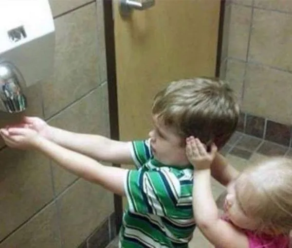 Dve deti v kúpeľni, jedno suší ruky pod sušičom, zatiaľ čo druhé si drží uši pri hluku.