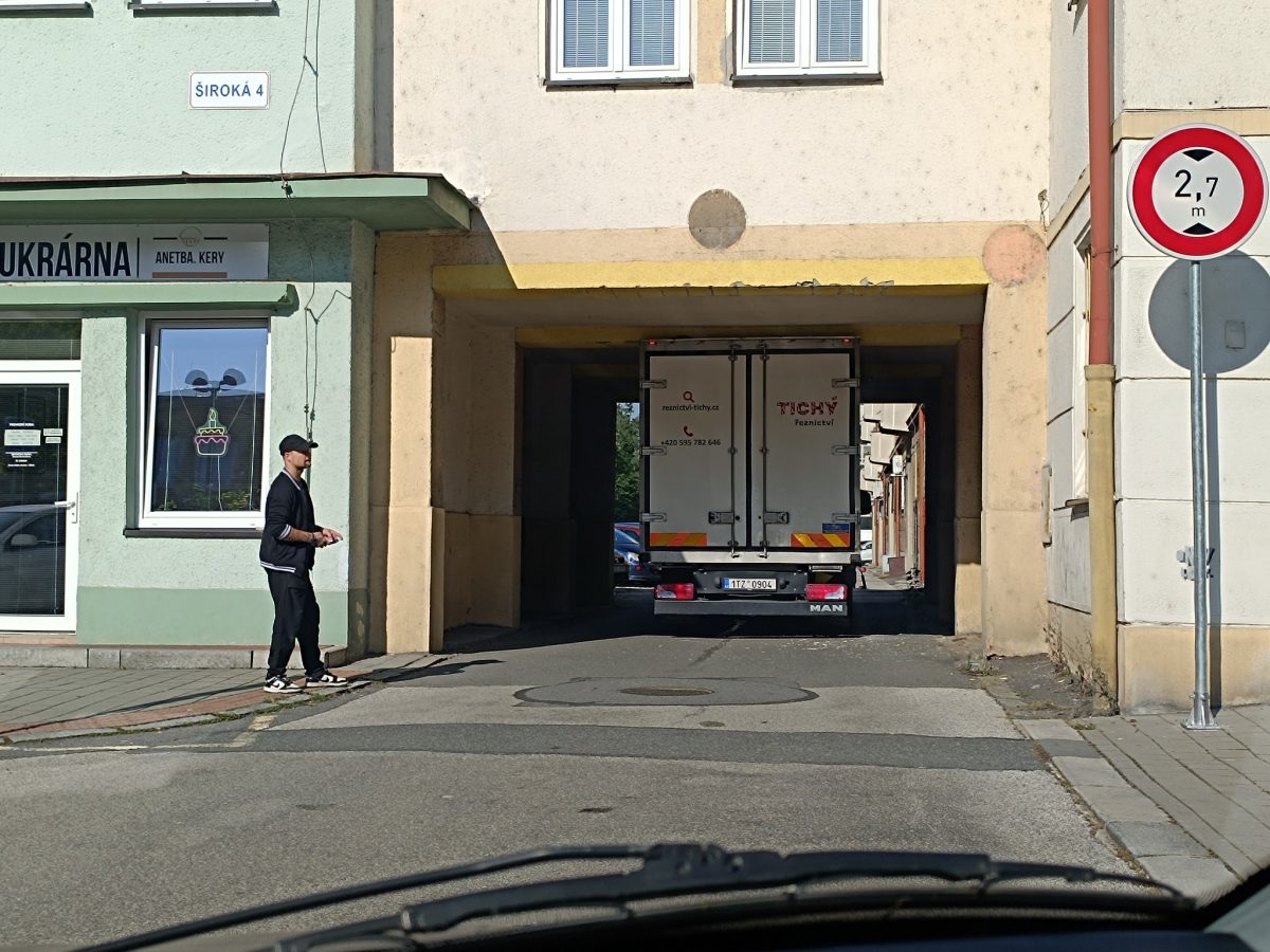 Kamión prechádzajúci úzkym priechodom pri cukrárni na Širokej ulici, vedľa prechádza chodec.