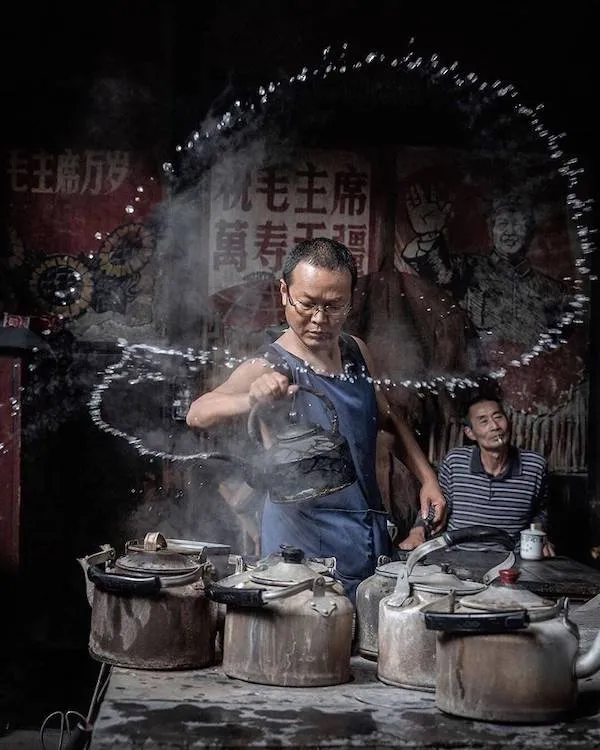 Muž v modrej zástere nalieva vodu z kanvice s pôsobivým oblúkom pary, zatiaľ čo druhý muž pozoruje.