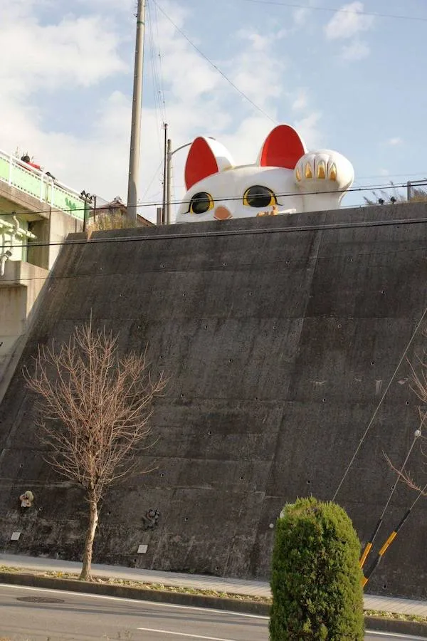 Obrázok mačky za múrom, vidieť len uši a oči, slnečný deň a cesta popri stene.