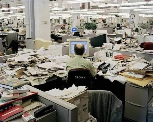 Kancelária plná papierov a neporiadku s mužom pri počítači uprostred pracovného chaosu.