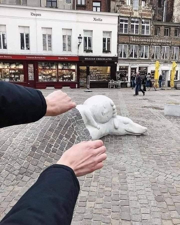 Osoba drží optický iluzórny panel na ulici s ležiacou sochou v pozadí.