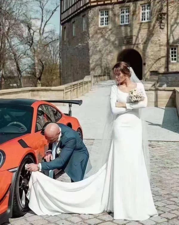 Ženích v obleku opravuje koleso červeného auta, nevesta v šatách sleduje situáciu pred historickou budovou.