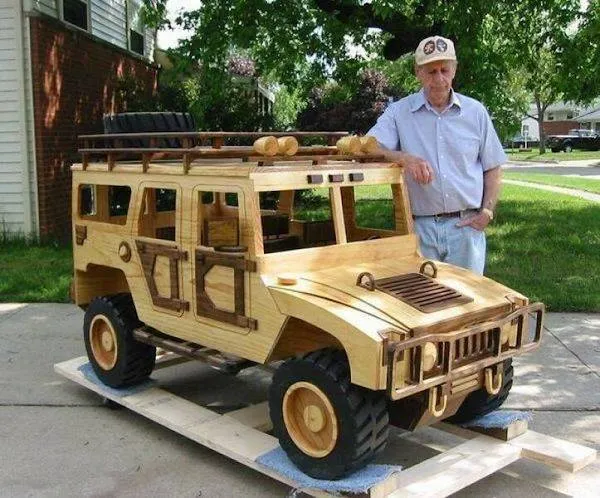 Muž stojí vedľa ručne vyrobeného dreveného modelu auta na dvore.