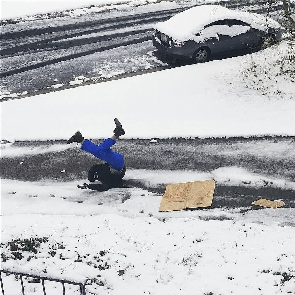 Osoba vtipne padá na zľadovatenej ceste počas zasneženého dňa, vedľa ležia kartónové dosky.