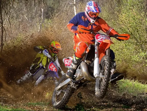 Motokrosoví jazdci pretekajú cez lesnú trať, zdolávajú blato a zákruty s veľkou rýchlosťou a sústredením.