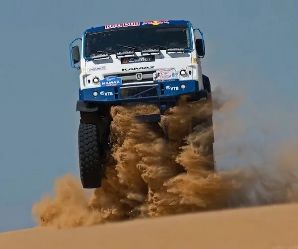 Kamión skáče cez piesočnú dunu na pretekoch rally, víri piesok za sebou.