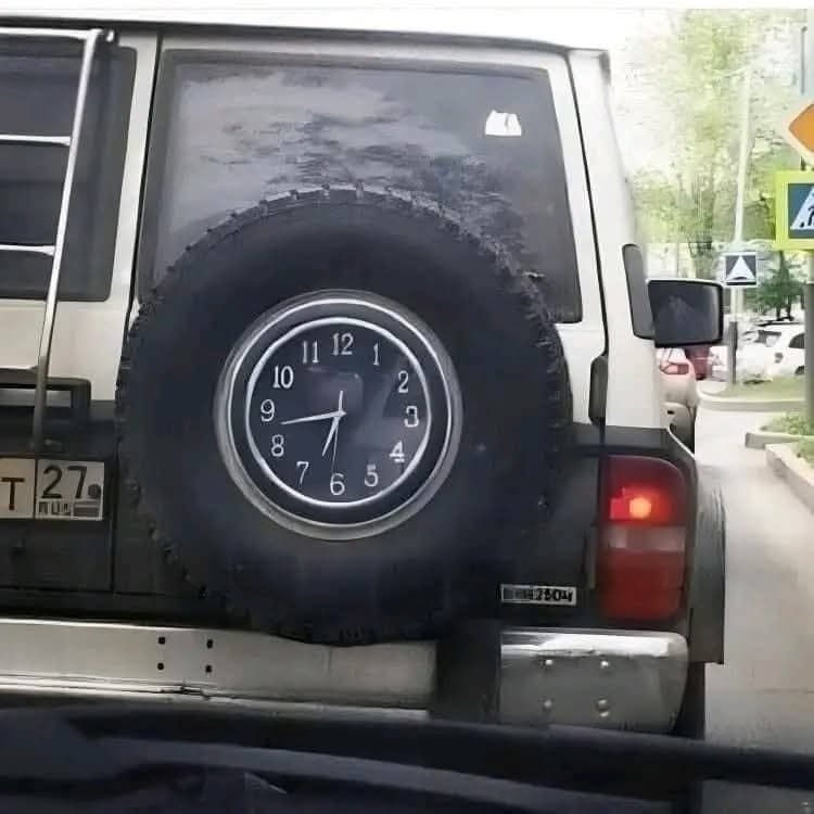 Rezervné koleso na vozidle s ciferníkom hodín namiesto centra, zaparkované na ulici pri zelených stromoch.