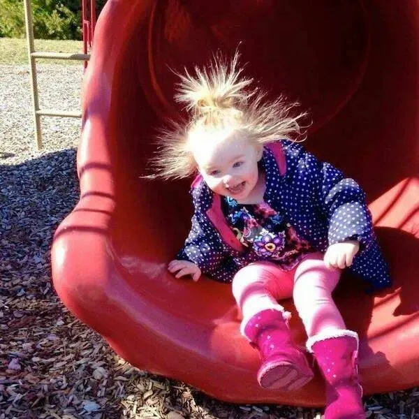 Dieťa na šmykľavke s vlasmi postavenými statickou elektrinou, oblečené v bunde a ružových topánkach.