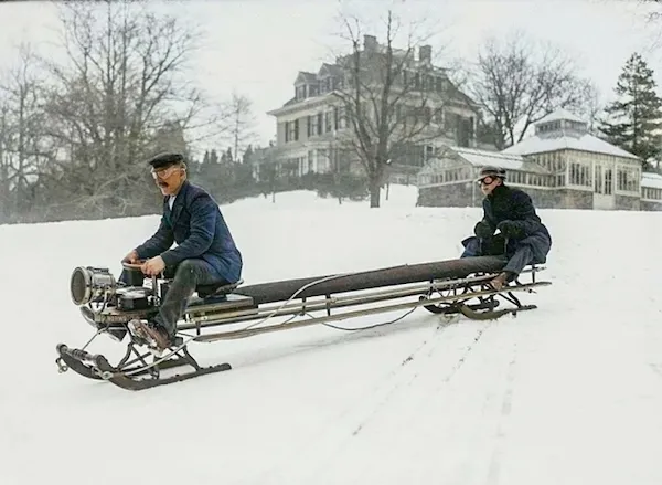 Dvojica na vintage motorových saniach na zasneženom svahu pred veľkým vidieckym domom.