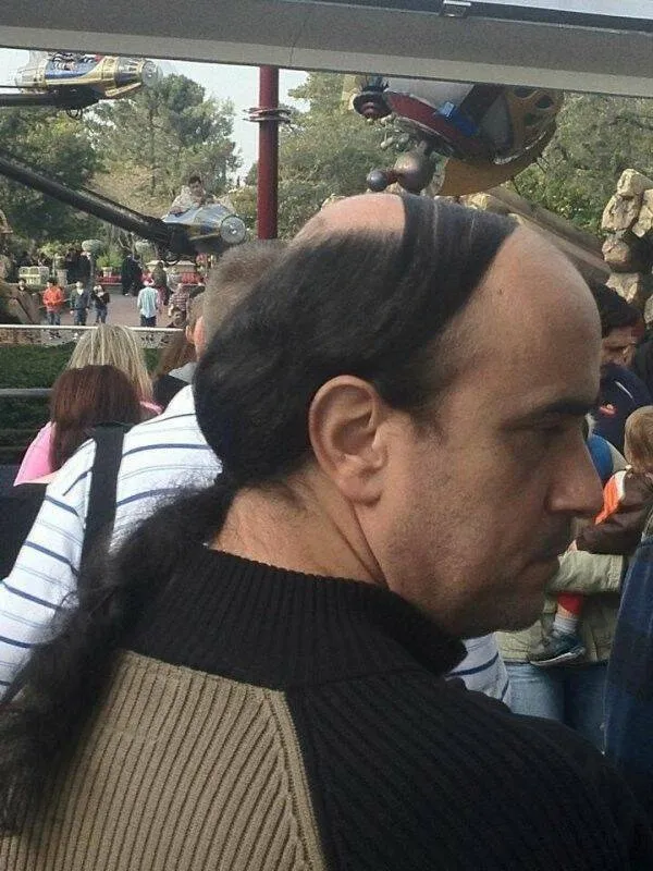 Muž s jedinečným účesom v zábavnom parku, pozadie s kolotočmi a ľuďmi.