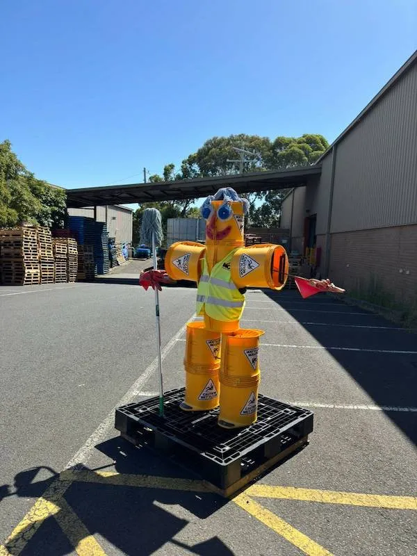 Figúrka z plastových sudov na palete v továrenskom areáli za slnečného dňa.