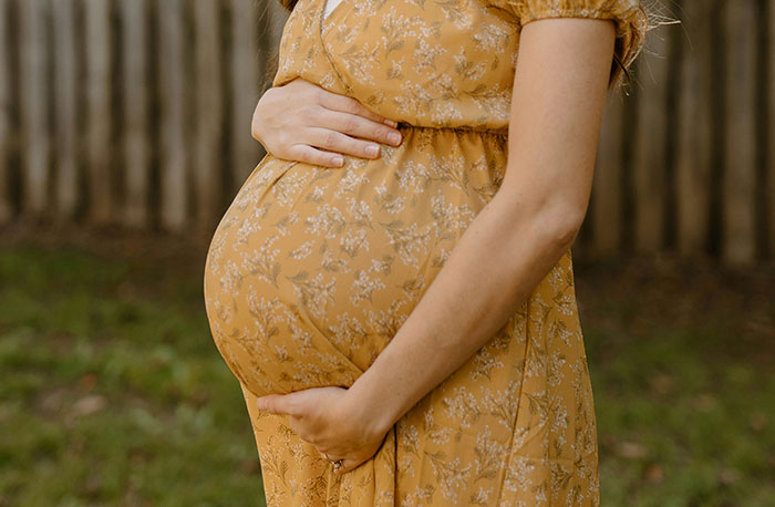 Tehotná žena v žltej kvetinovej šatách si drží brucho na zelenom pozadí.