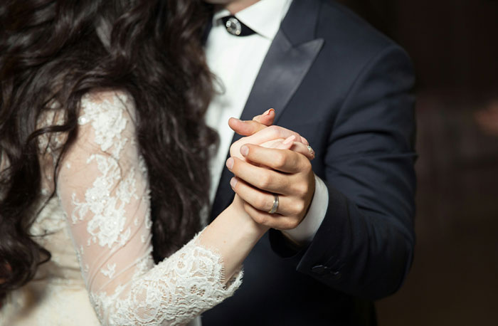 Pár držiaci sa za ruky počas tanca na svadbe, nevesta v čipkovaných šatách, ženích v tmavom obleku.