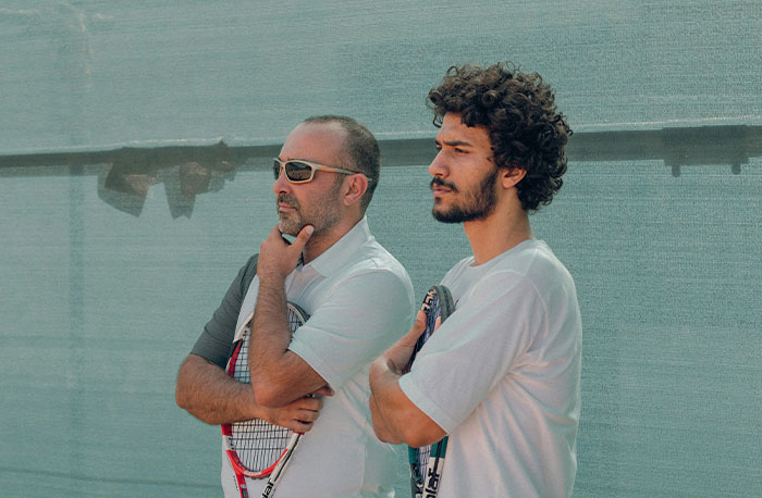 Dvaja tenisti uvažujú, držiac rakety, pri pohľade na kurt v slnečných okuliaroch a športovom oblečení.