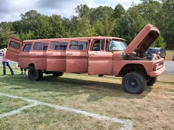 Dlhá ružová dodávka s otvorenou kapotou a dverami na tráve, obklopená lesom.
