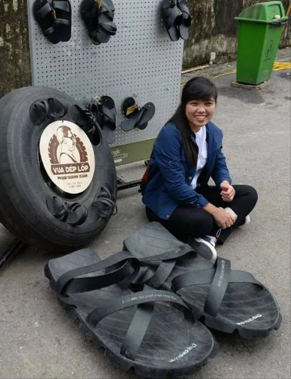Žena sediaca vedľa obrovských čiernych sandálov vyrobených z pneumatík, s panelom a prevádzkou v pozadí.
