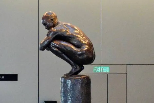 Socha človeka v podrepe na stĺpe v galérii, moderný umelecký štýl. Výstražné svetlo vystupu v pozadí.