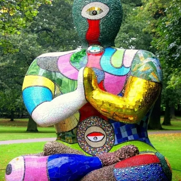 Farebná socha postavy s mozaikovou textúrou, sediaca v parku s rukami v meditačnej póze.