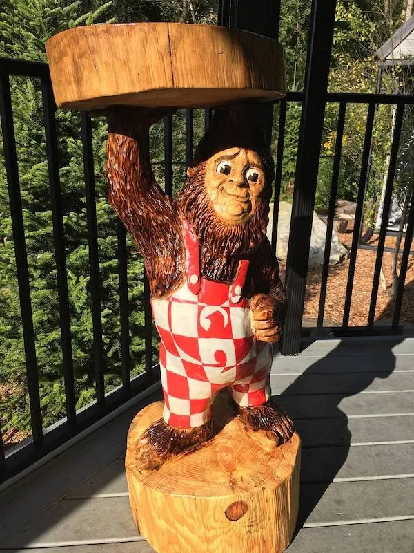 Drevená socha opice v kockovaných nohaviciach držiaca tanier na terase, obklopená stromami.