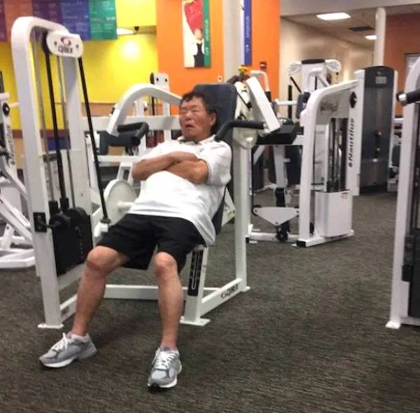 Osoba oddychuje na posilňovacom stroji v posilňovni, oblečená v bielom tričku a čiernych šortkách.