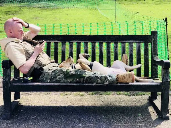 Muž a pes relaxujú na lavičke v parku, muž pozerá do mobilu a pes leží pohodlne na chrbte.