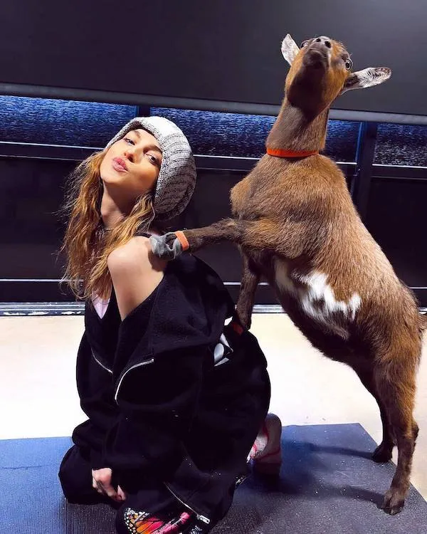 Osoba pózuje s kozou na čiernom pozadí, kozia labka na pleci, zábavná momentka v interiéri.