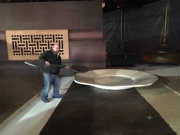 Muž drží veľkú lyžicu vedľa obrovskej taniere v tmavej miestnosti.