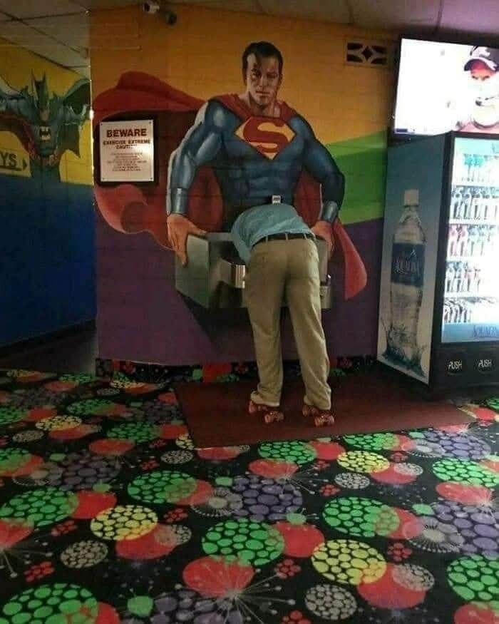 Muž pije vodu z fontány pod stenom vymaľovaným Supermanom na farebnej kobercovej podlahe.