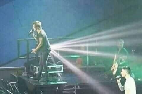 Spevák na pódiu s dramatickým svetelným efektom, pri čom lúče pôsobia, akoby vychádzali z jeho chrbta.