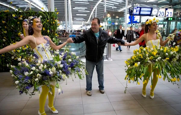 Osoby v kvetinových kostýmoch tancujú na letisku, držia muža za ruky a usmievajú sa.
