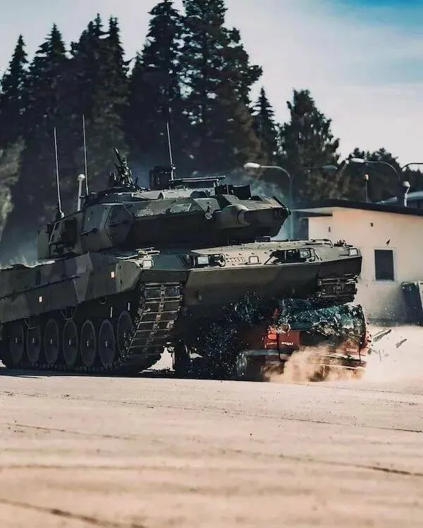 Tank prechádza cez auto na vojenskom cvičení, okolie sú stromy a budovy v pozadí.