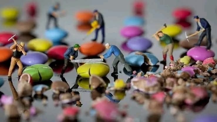 Miniatúrni robotníci pracujú s farebnými sladkosťami ako obrovskými balvanmi na zrkadlovej ploche.