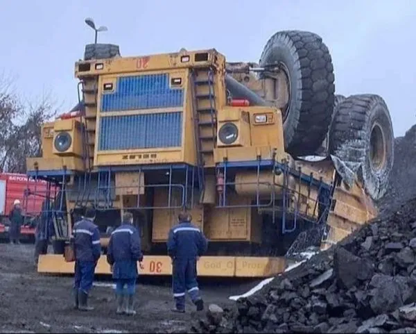 Obrátený ťažký nákladný automobil s tromi pracovníkmi sledujúcimi situáciu na stavenisku plnom kameniva.