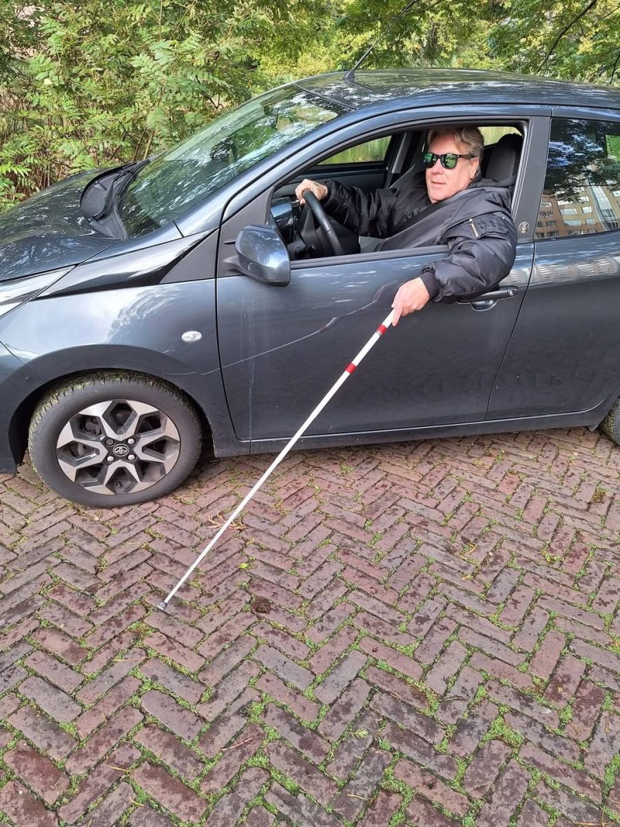 Muž v aute s bielou slepeckou palicou, parkovanie na dláždenej ceste, okolo sú stromy.